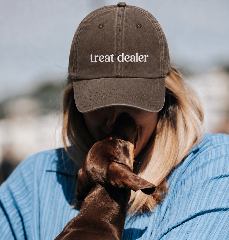 Woman wearing a brown "treat dealer" cap being nuzzled by a dachshund.