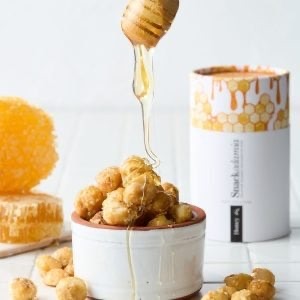 Honey drizzling over roasted macadamias beside a honeycomb Snackadamia canister.